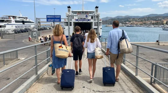 Eine vierköpfige Familie von hinten fotografiert, die mit Strandtaschen und Rollkoffer auf eine moderne Fährampe in einem mediterranen Hafen zugeht
